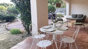 An outdoor patio with white metal chairs and a table set for dining, surrounded by a garden and a vehicle in the background. - A haven of peace. Ground floor of a villa near the harbor with a private garden and entrance (Sanary-sur-Mer)