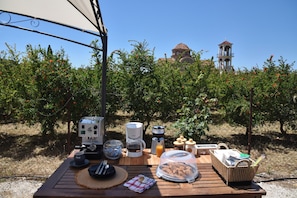 A picnic setup with a coffee maker, a basket, and a table in an outdoor setting with a view of a building and trees.
