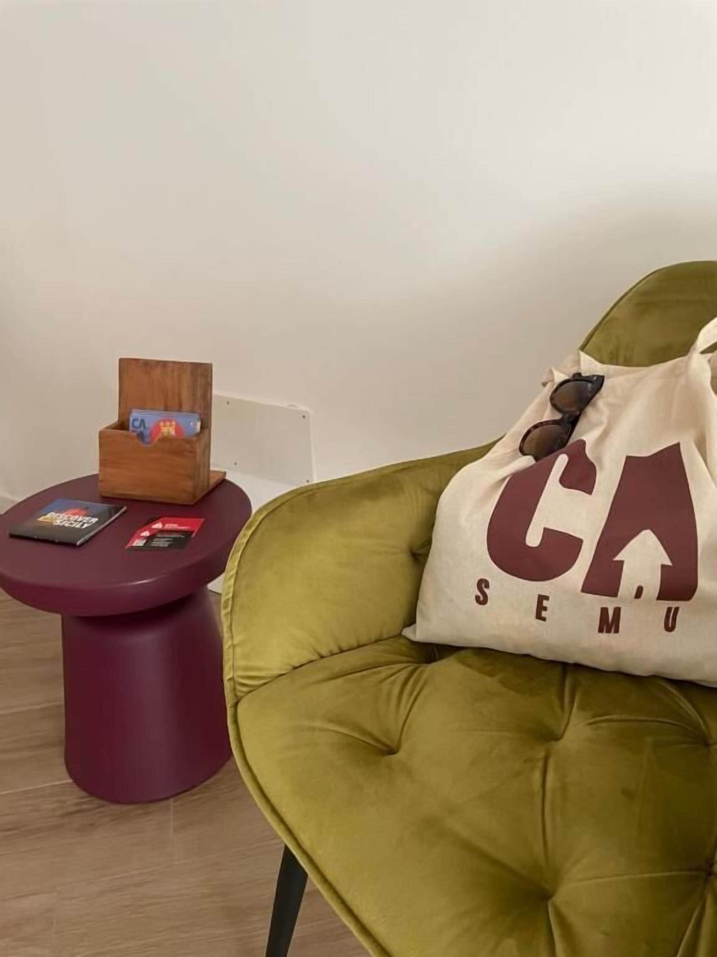 A green chair with a cushion, a small round table with a magazine holder, and a wall-mounted shelf with a book.