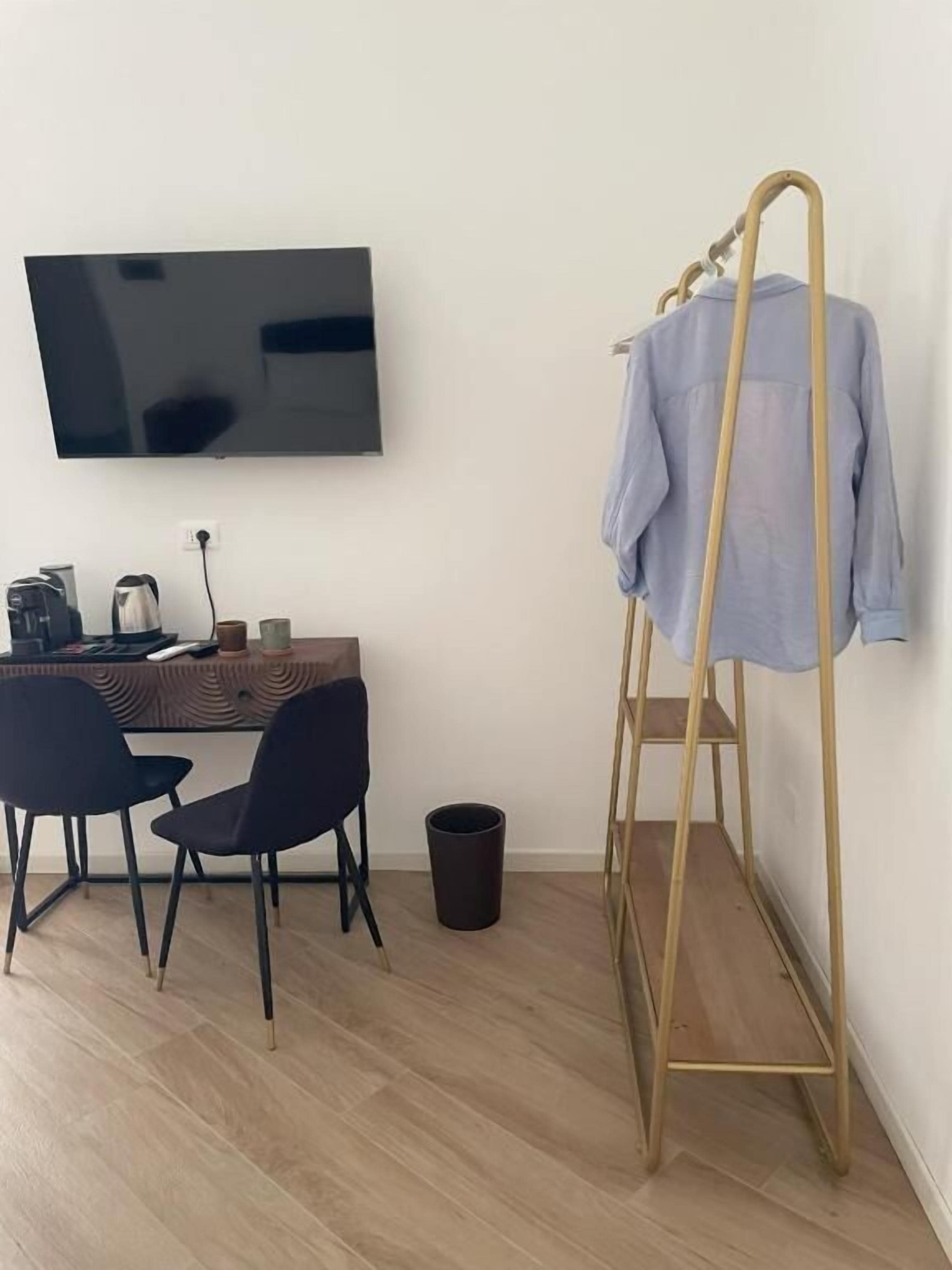 A room with a TV mounted on the wall, a desk with chairs, a coat rack, and a trash bin.