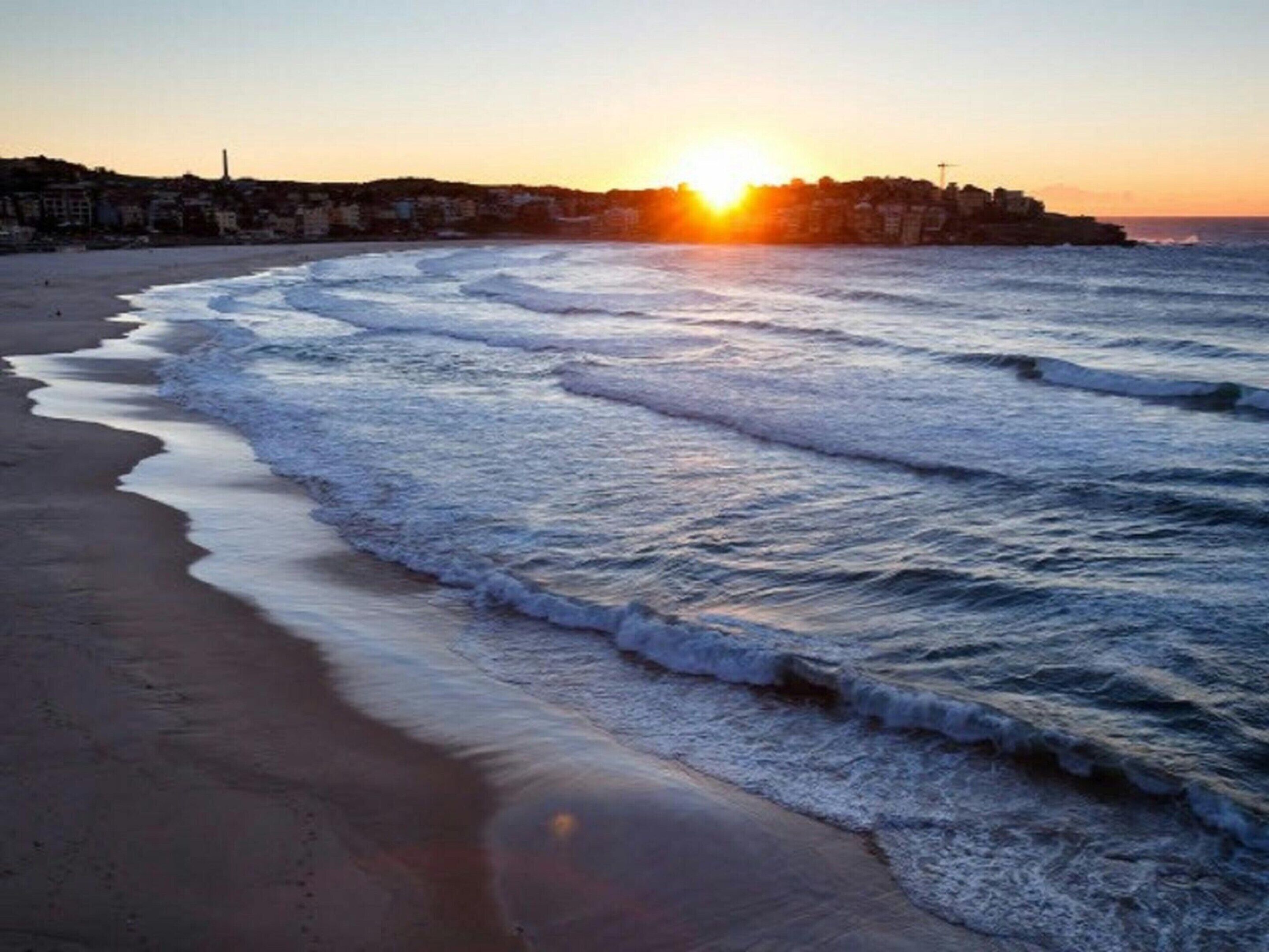 Une plage avec des vagues, un coucher de soleil et des bâtiments en arrière-plan.