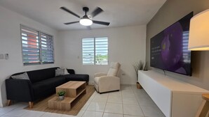 Una sala de estar moderna con un sofá negro, un sillón color beige, una mesa de centro de madera, un ventilador de techo y un televisor de pantalla plana grande montado en la pared.
