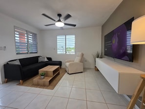 Una sala de estar moderna con un sofá negro, un sillón color beige, una mesa de centro de madera, un ventilador de techo y un televisor de pantalla plana grande montado en la pared.
