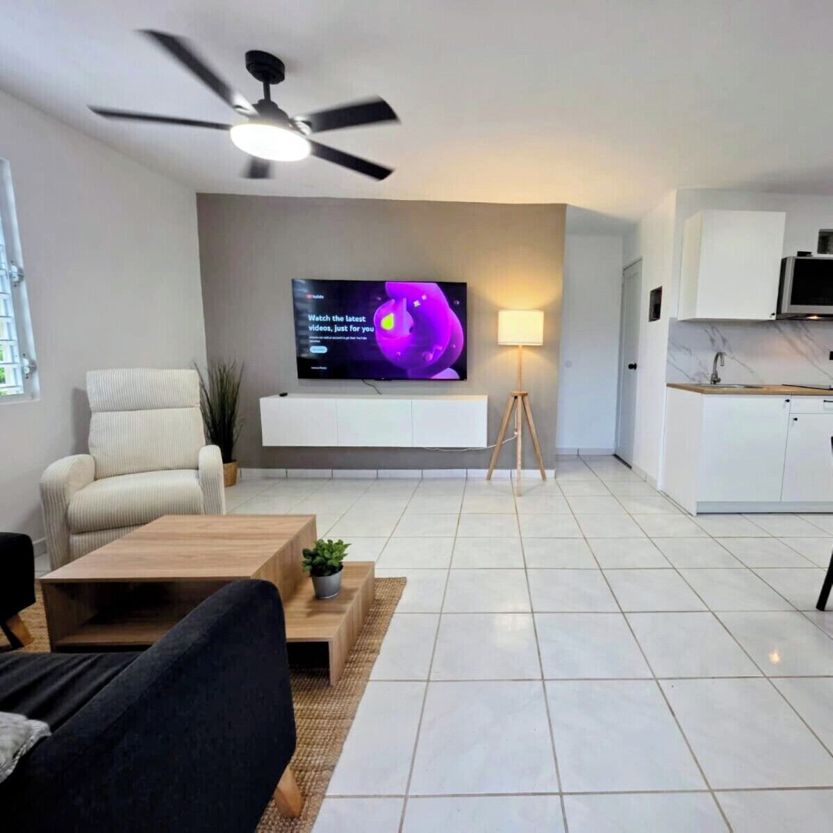 A modern living room with a flat-screen TV, a ceiling fan, a sofa, a coffee table, and a kitchen area with cabinets and a countertop.