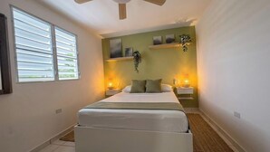 Un dormitorio con cama, ventilador de techo, ventana con persianas y estantes empotrados con objetos decorativos.