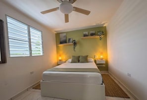 Un dormitorio con cama, ventilador de techo, ventana con persianas y estantes empotrados con objetos decorativos.