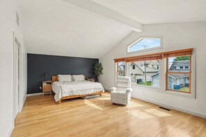 Un dormitorio con cama, mesita de noche, silla y ventanas amplias.