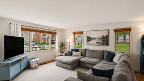 Ett vardagsrum med en hörnsoffa, en platt-TV, ett litet bord med en växt och stora fönster med utsikt över ett hus och träd.