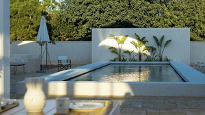 Uma área de piscina ao ar livre moderna com design minimalista, incluindo um espaço de estar com cadeiras e uma mesa, cercada por vegetação exuberante e uma parede branca.