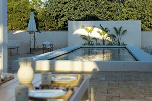 Un'area piscina moderna all'aperto, dal design minimalista, con zona relax con sedie e tavolo, immersa nel verde e delimitata da un muro bianco.