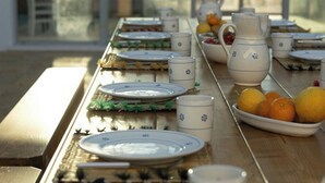Mesa de jantar posta com pratos, copos e jarra, com vista para um edifício através de janelas amplas.