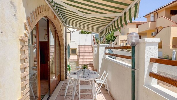 En overdækket terrasse med et bord og stole, et træhegn og en bygning i baggrunden.