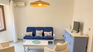 En stue med en blå sofa, hvide lænestole, et sofabord, et fjernsyn på et skab og en hængende lampe.