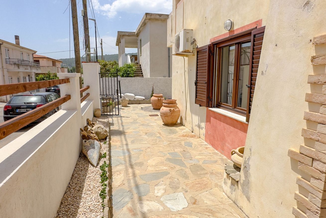 A narrow stone-paved courtyard with terracotta pots, a wooden fence, and a building with a red window frame.