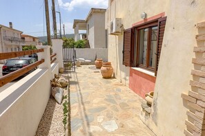 A narrow stone-paved courtyard with terracotta pots, a wooden fence, and a building with a red window frame. - Appartamento l'Anfora (Cala Gonone)