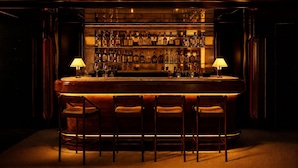 Un bar con mostrador de madera, varias botellas de licor y cuatro taburetes.
