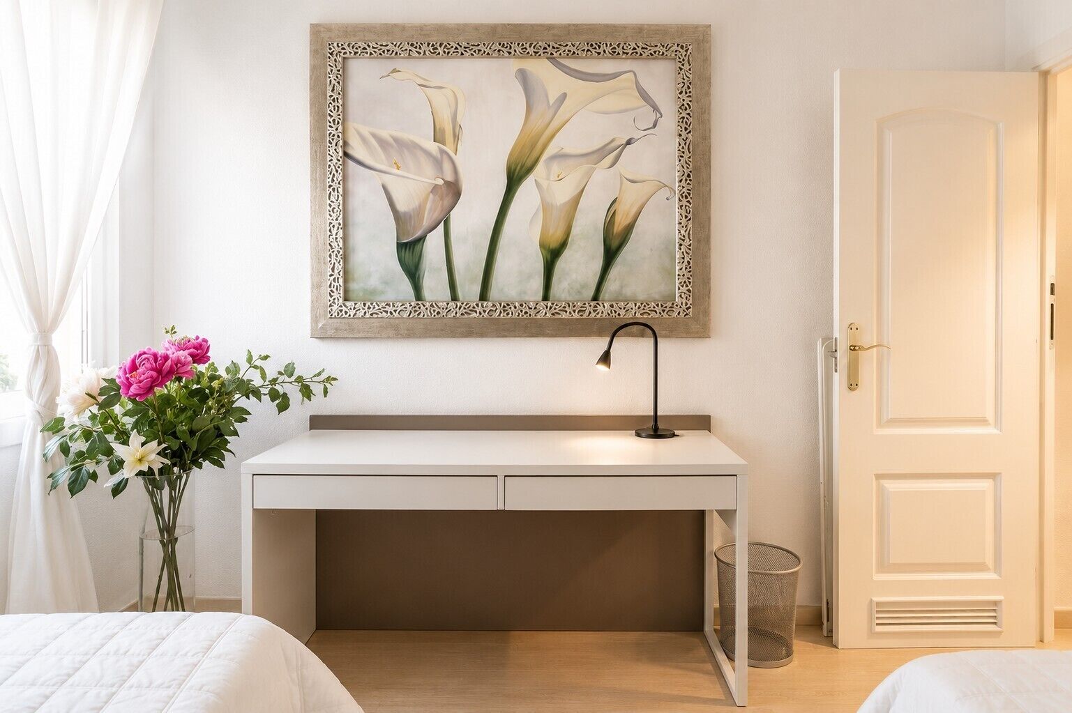 A hotel room with a bed, a bedside table with a lamp, a desk with a chair, a door, and a framed artwork on the wall.