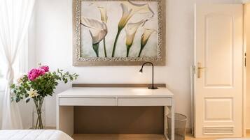 A hotel room with a bed, a bedside table with a lamp, a desk with a chair, a door, and a framed artwork on the wall.