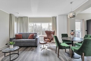 A modern living room with a grey sofa, a round dining table with green chairs, a brown chair, and a small side table with a plant. - ALVA Apartments Snorrabraut (Reykjavík)