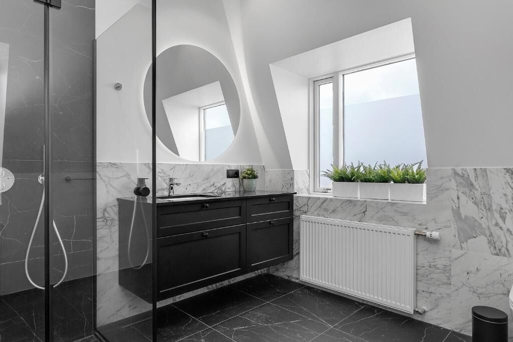 A modern bathroom with a glass shower enclosure, a large mirror, a black vanity, a white radiator, and a window with a plant.