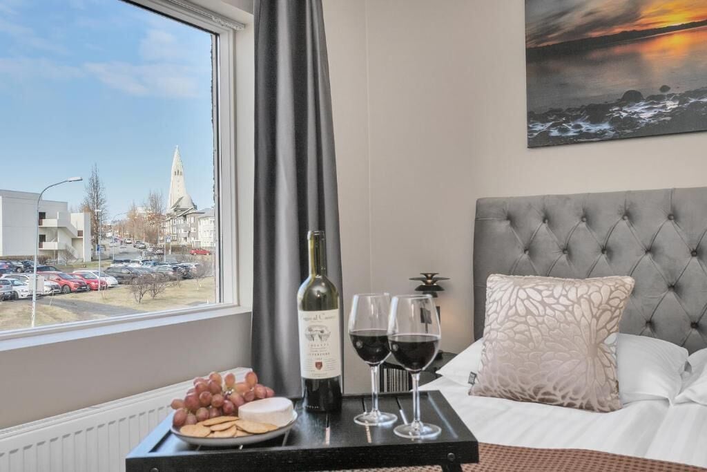 A hotel room with a bed, a window with a view of buildings and cars, a bottle of wine, glasses, grapes, and cheese on a tray.