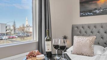 A hotel room with a bed, a window with a view of buildings and cars, a bottle of wine, glasses, grapes, and cheese on a tray.