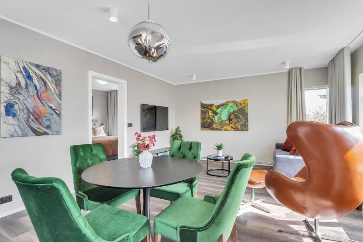 Uma sala de estar moderna com uma mesa redonda, poltronas estofadas em verde, uma cadeira marrom singular, uma pintura abstrata grande e uma televisão.
