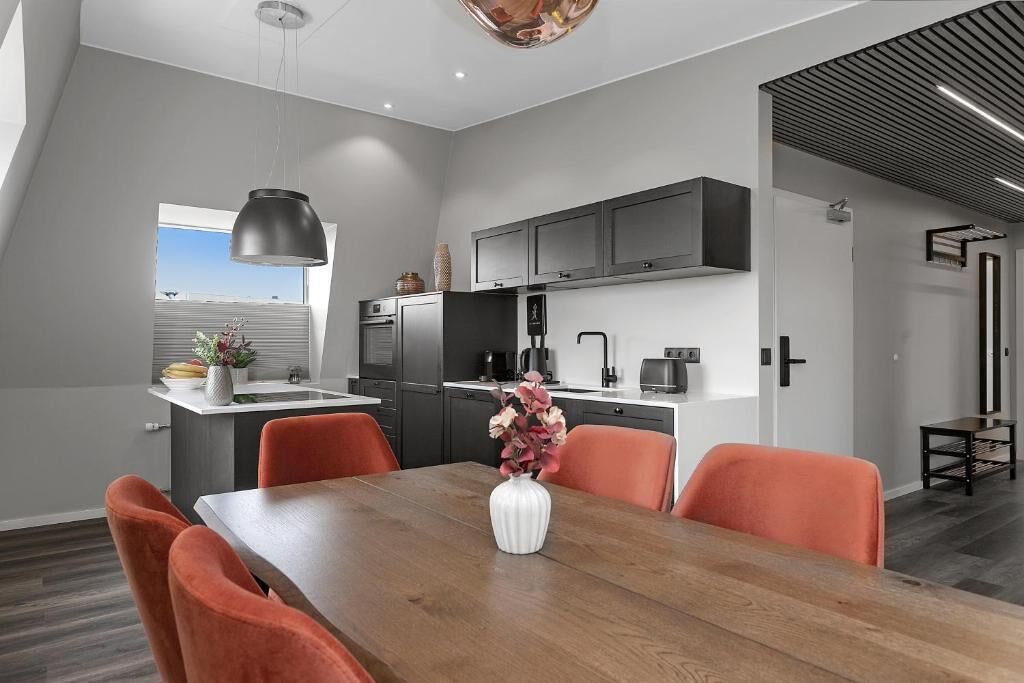 A modern kitchen with dark cabinets, a dining table with orange chairs, and a vase with flowers.