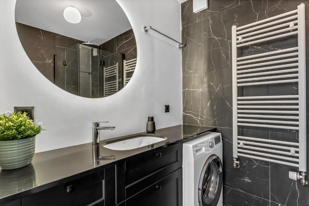 Un baño moderno con un espejo redondo, un lavabo, una lavadora y un perchero para toallas.