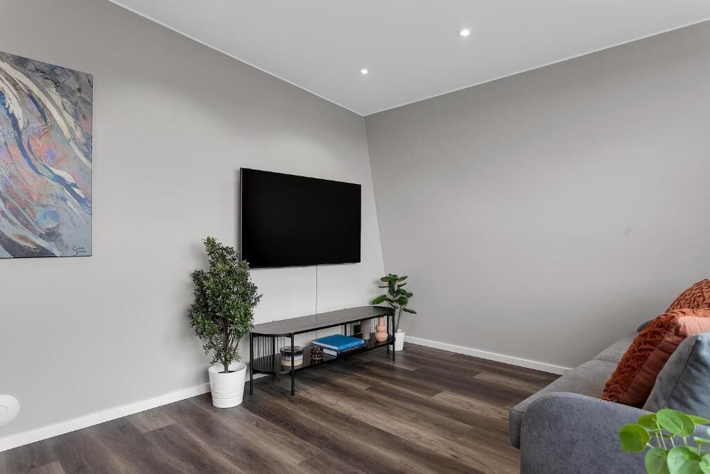 A modern living room with a flat-screen TV mounted on the wall, a black console table with decorative items, a grey sofa, and a large abstract painting on the left wall.