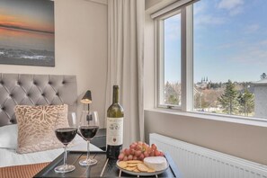 Ett hotellrum med en säng, utsikt över staden, en flaska vin, glas och en tallrik med vindruvor och kex.