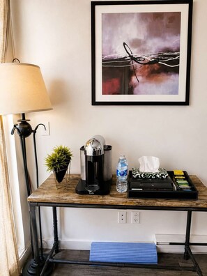 Una habitación con escritorio, lámpara, cafetera, botella de agua, pañuelos de papel y un cuadro abstracto enmarcado.