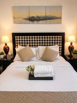 A neatly made bed with a checkered headboard, two bedside lamps, a cityscape painting above, and a tray with a towel and flowers. - Cozy 2 BR-5min Downtown • Walk to Shops & Transit (Vancouver)