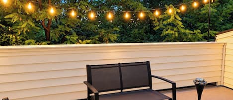 A patio with a bench and a glass table, string lights, and a white wall.