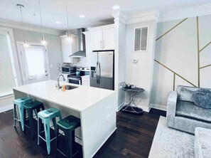 Een moderne keuken met witte kasten, een centraal kookeiland met barstoelen, roestvrijstalen apparatuur, en een zithoek met een grijze bank.