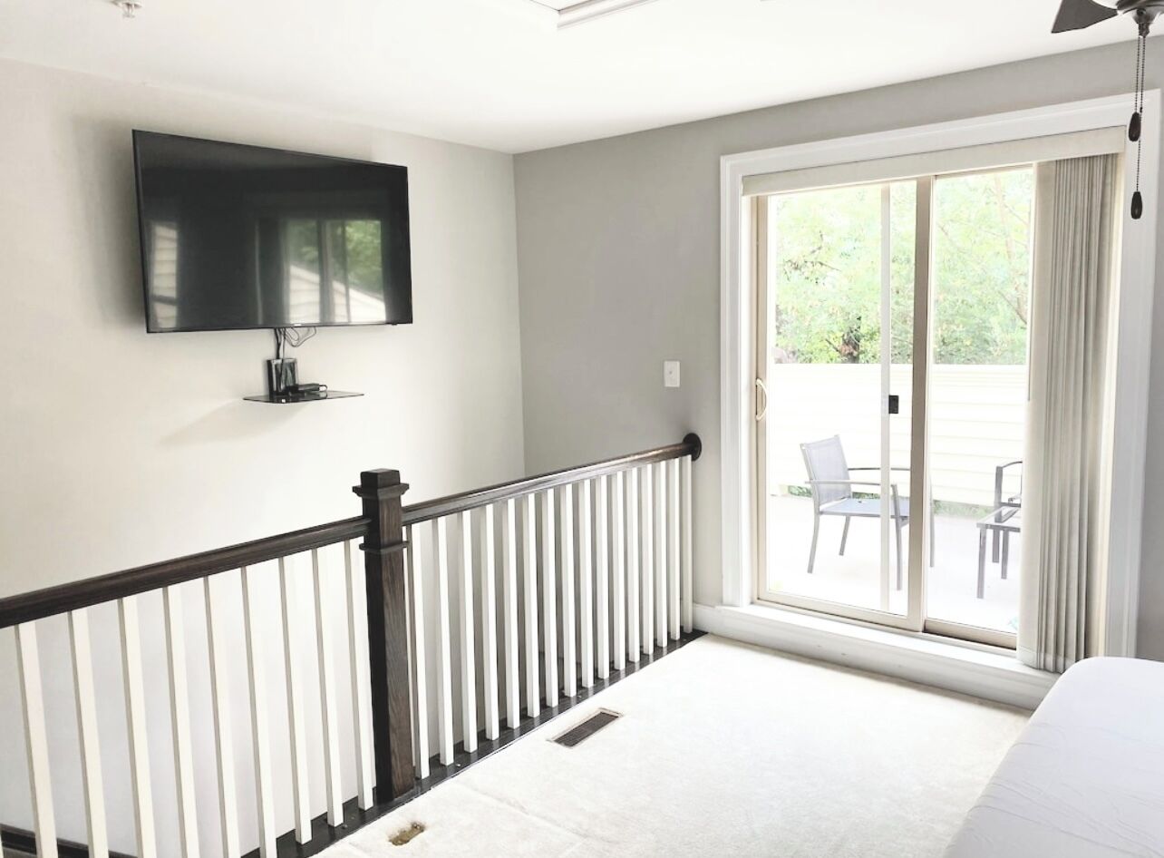 A room with a flat-screen TV mounted on the wall, a balcony with a view of outdoor furniture, and a white floor.