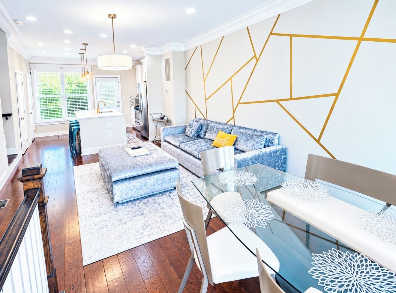 A modern living room with a glass dining table, blue patterned sofas, and a geometric wall design.