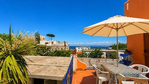 A terrace with a table and chairs, a large umbrella, and a view of the sea and buildings. - Hotel Aragonese Ischia (Ischia)