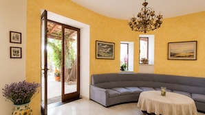Un salón con un sofá gris, una mesa redonda con mantel de encaje, una araña y una puerta que da al exterior.