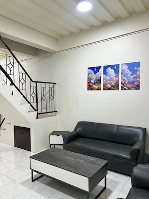 A modern living room with a black leather sofa, a grey coffee table, a white side table, and a staircase with black railings. - Jaz Urban Stay - 337f (Angeles City)