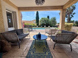 Eine Terrasse im Freien mit Korbmöbeln, einem gemusterten Teppich und Blick auf Pool und Grünflächen.