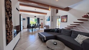 Ein modernes Wohnzimmer mit einer Couch, einem Couchtisch und einem Kamin. Es gibt eine Essecke mit Stühlen und einem Tisch. Der Raum verfügt über Holzbalken an der Decke und eine Steinwand.