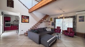 Ein modernes Wohnzimmer mit einem grauen Ecksofa, einer Holztreppe, einem Essbereich mit roten Stühlen und einer Küche im Hintergrund.
