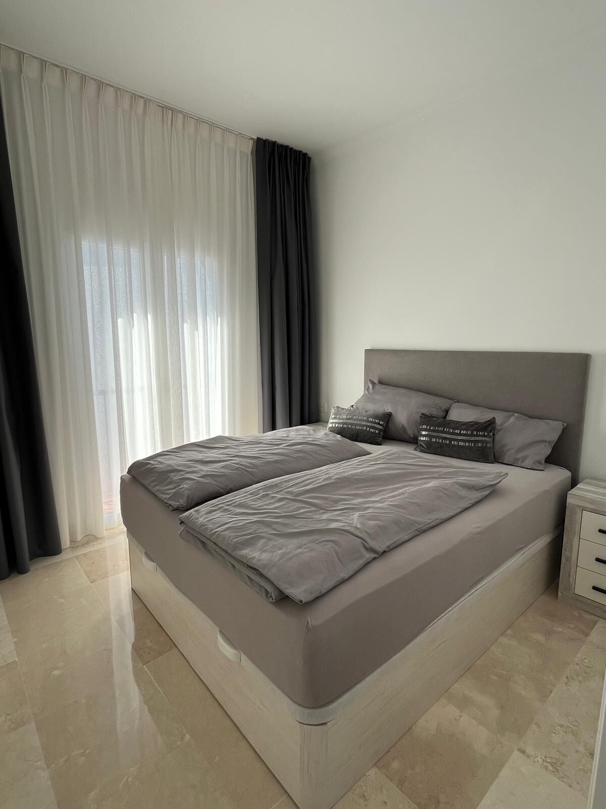 A modern bedroom with a large bed, a nightstand, and a window with curtains.