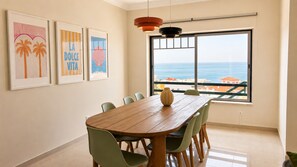 Un comedor con mesa y sillas de madera, obras de arte en la pared, un ventanal con vistas al mar e iluminación colgante.