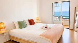 Un dormitorio con cama, almohadas, toalla, mesita de noche, lámpara y vista al océano.