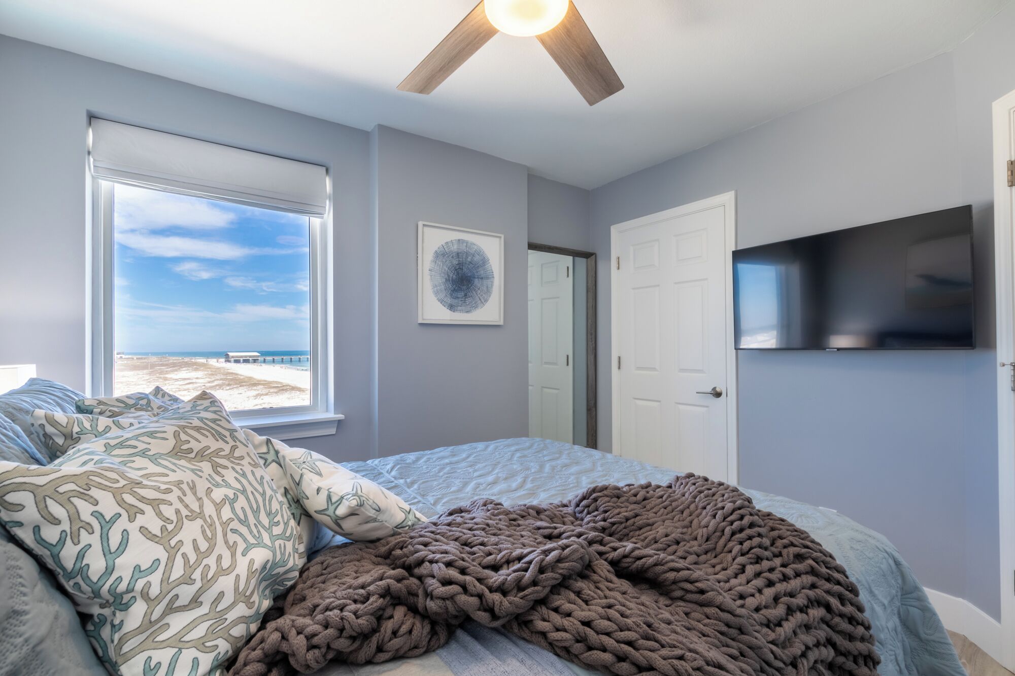 Une chambre avec un lit, une grande fenêtre donnant sur la plage, un ventilateur de plafond et un téléviseur à écran plat fixé au mur.