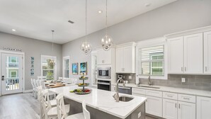 A modern kitchen with white cabinetry, a central island, and a dining area with white chairs. - Sea Clearly | Close to Beach | Golf Cart (LSV) | 4 Bikes | Pool (Santa Rosa Beach)
