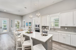 A modern kitchen with white cabinetry, a central island, and a dining area with white chairs. - Sea Clearly | Close to Beach | Golf Cart (LSV) | 4 Bikes | Pool (Santa Rosa Beach)