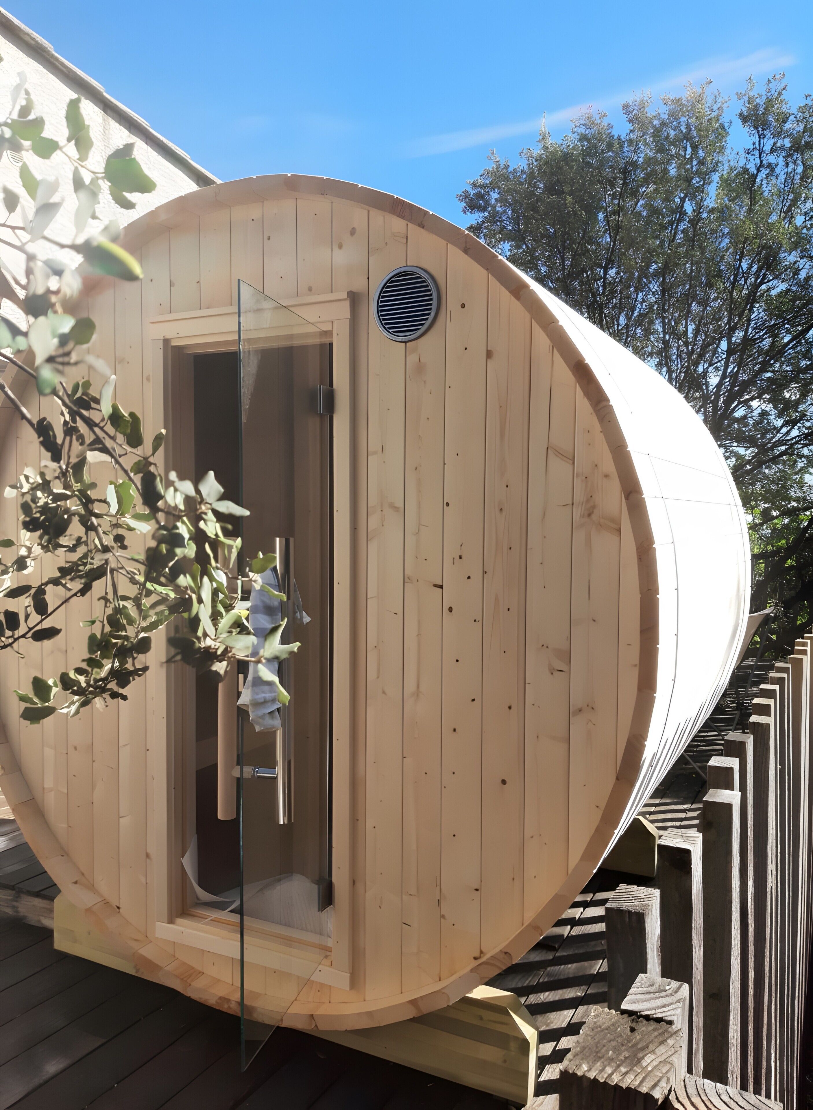 A wooden sauna with a glass door, a ventilation vent, and a wooden deck surrounding it.